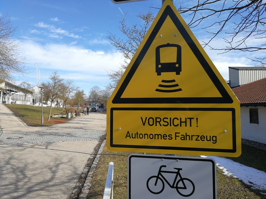 Ein Warnschild "Vorsicht! Autonomes Fahrzeug". Im Hintergrund fährt das besagte Fahrzeug.